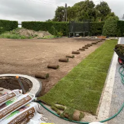 Układanie trawy z rolki w ogrodzie, widoczne rolki trawy, taczka z trawą, worki z korą ogrodniczą, przygotowany teren, żywopłot w tle.