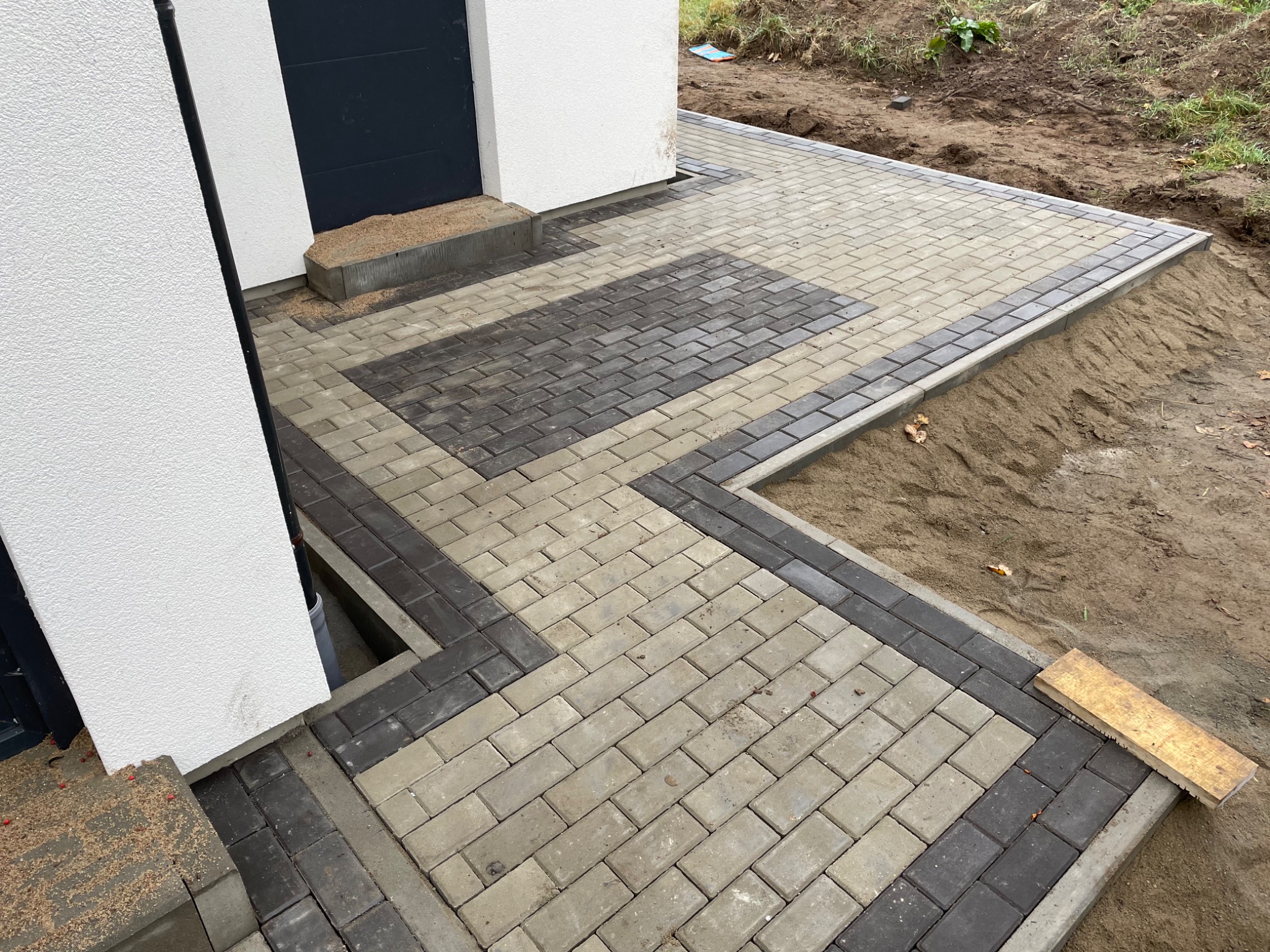 Ułożona kostka brukowa w dwóch odcieniach szarości tworząca wzór na podjeździe przed domem, widoczne niedokończone prace ziemne po bokach.