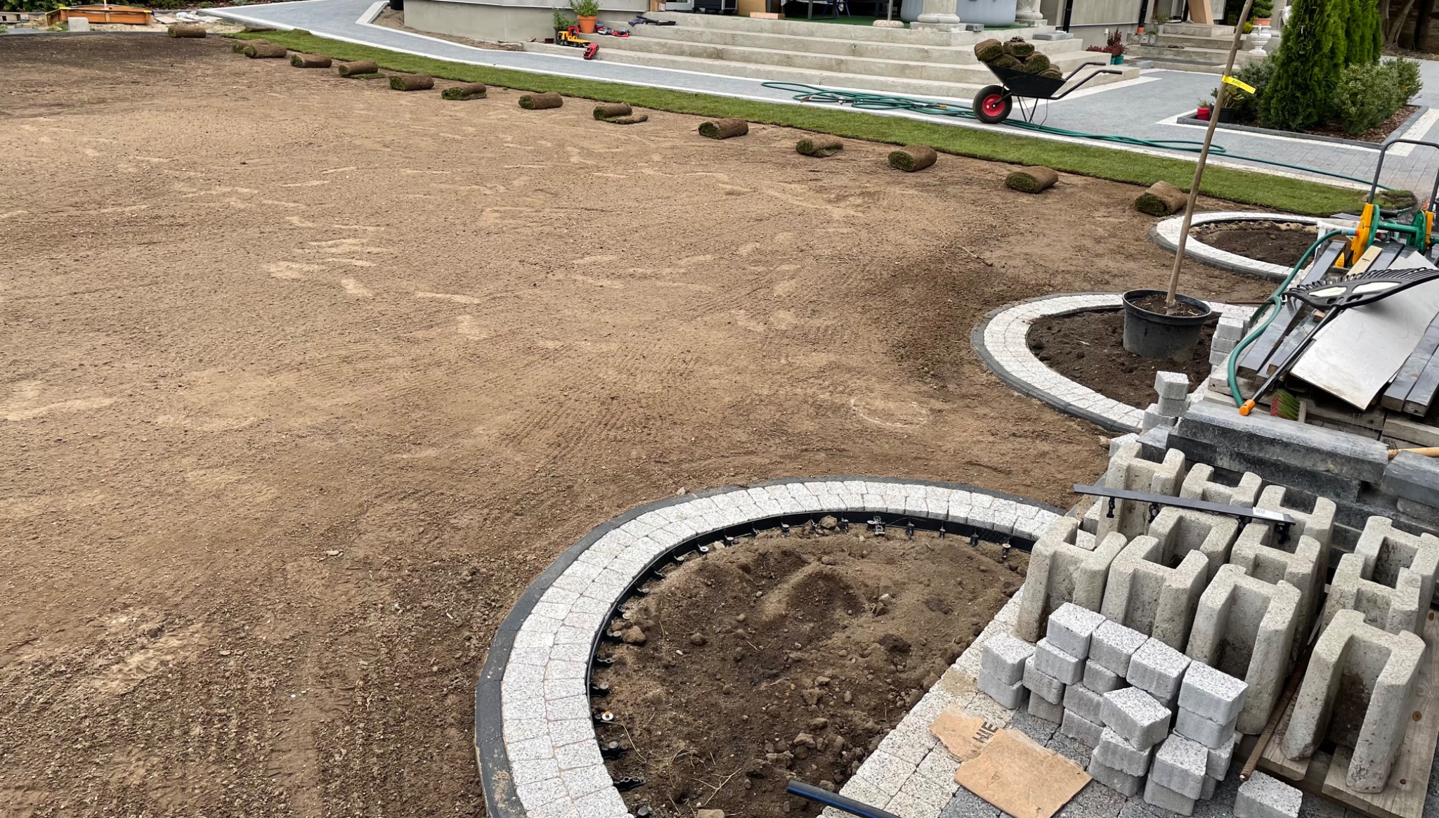 Przygotowany teren pod trawnik z rolki, widoczne ułożone rolki trawy, obrzeża rabat z kostki granitowej i elementy betonowe do budowy ścieżki ogrodowej