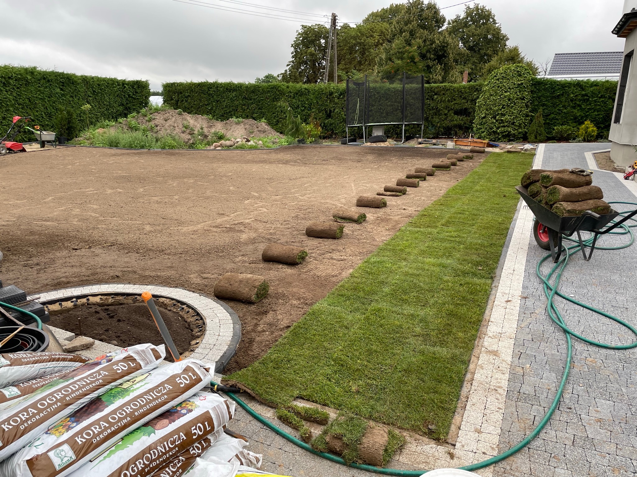 Układanie trawy z rolki w ogrodzie, widoczne rolki trawy, taczka z trawą, worki z korą ogrodniczą, przygotowany teren, żywopłot w tle.