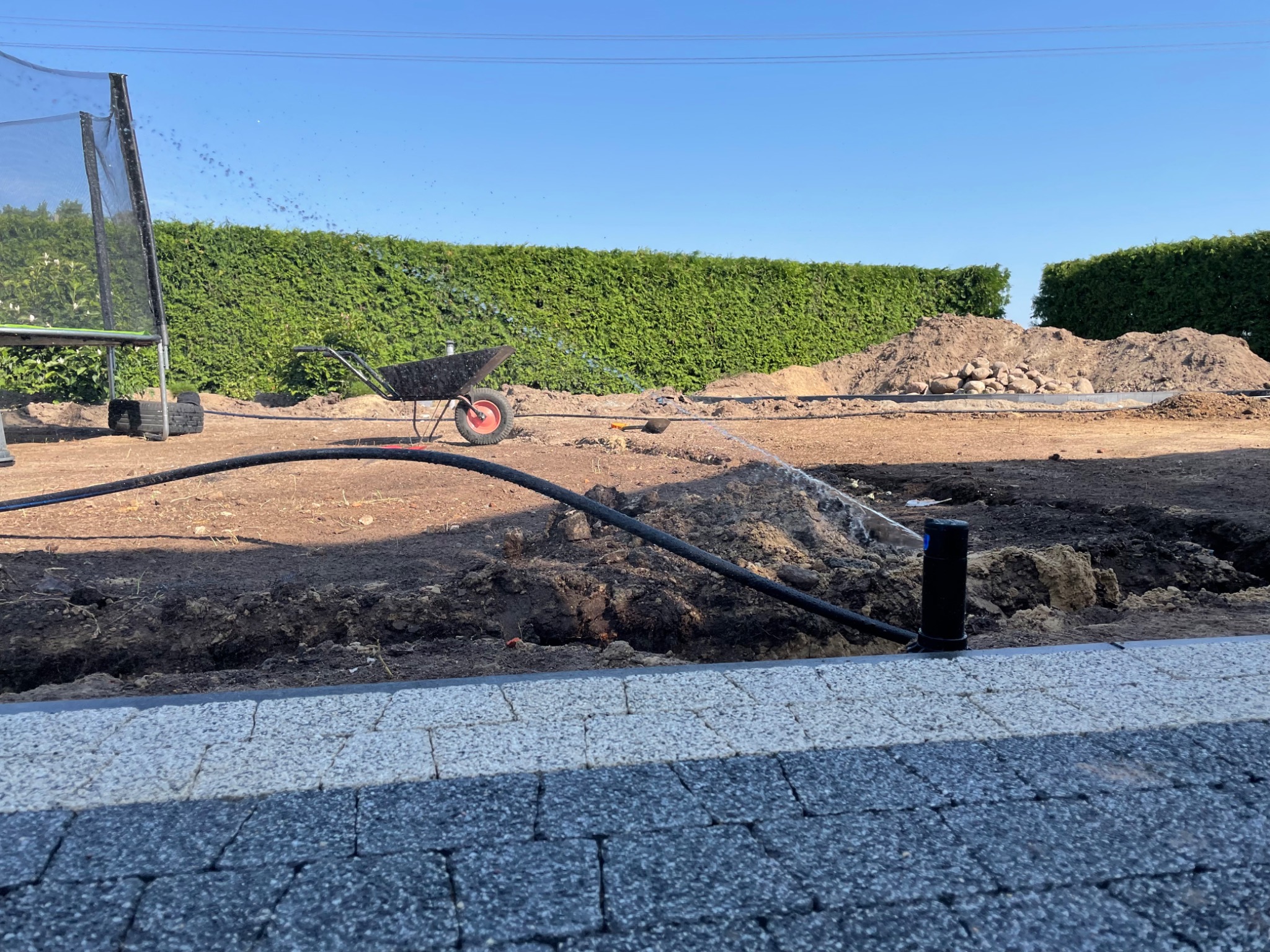 Instalacja systemu nawadniania w ogrodzie: widoczny fragment trawnika z wykopanym rowem, czarny przewód doprowadzający wodę do zraszacza, w tle taczka i zielony żywopłot.