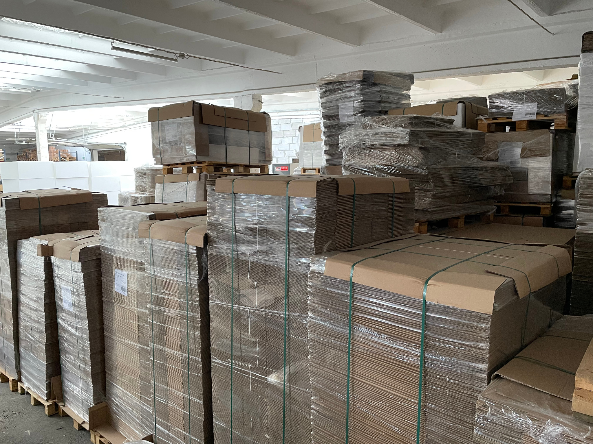 Magazyn z paletami wypełnionymi płaskimi, kartonowymi arkuszami, zabezpieczonymi folią i taśmą, pod białym, przemysłowym sufitem.