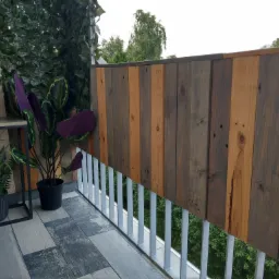 Balkon z drewnianą balustradą z desek o różnej kolorystyce, uzupełniony o rośliny w donicach i sztuczną zieloną ścianę.