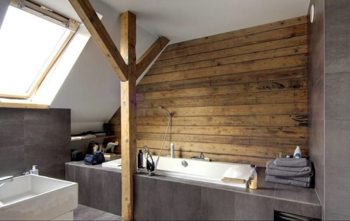 Nowoczesna łazienka na poddaszu z drewnianą ścianą i belką konstrukcyjną, białą wanną oraz oknem dachowym.