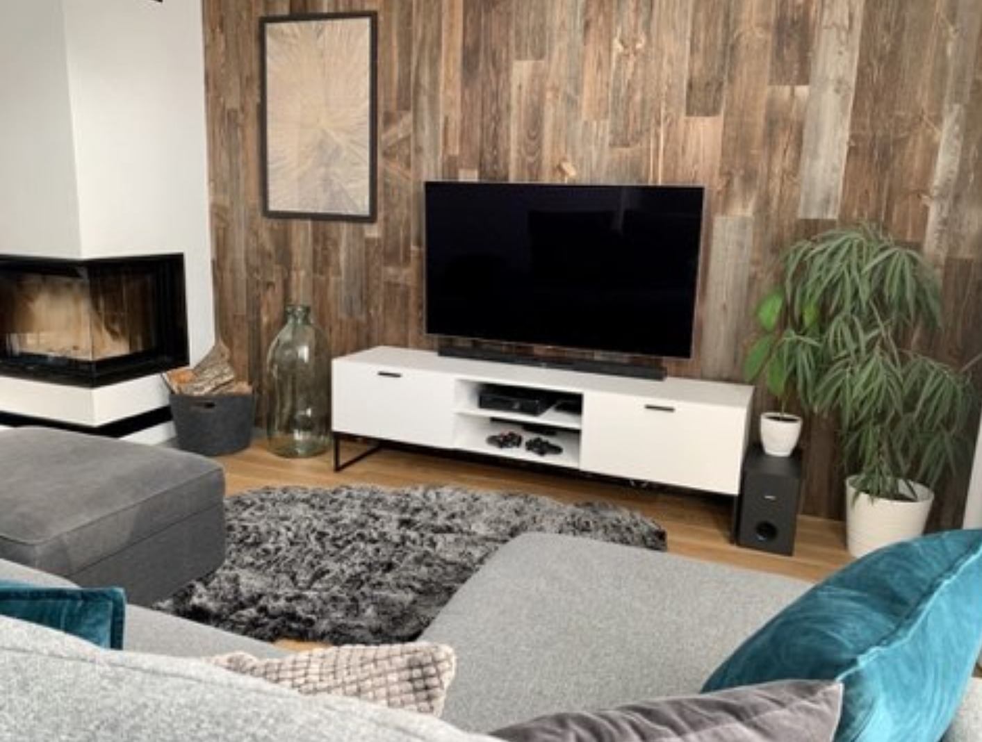 Nowoczesny salon z drewnianą ścianą, kominkiem narożnym, białym stolikiem RTV i szarą sofą modułową.