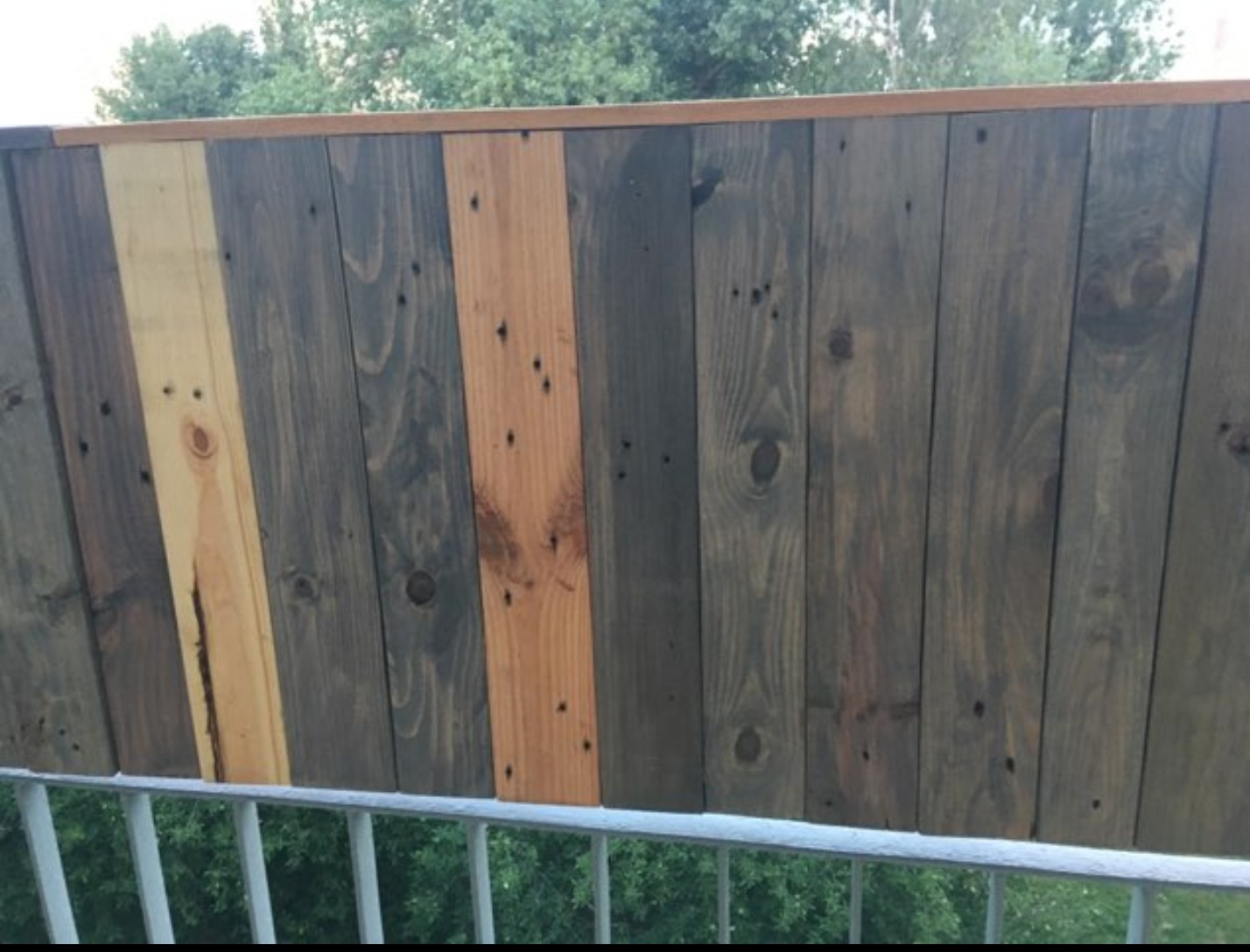 Fragment balustrady balkonowej z pionowo ułożonych desek w różnych odcieniach brązu, z widocznymi sękami i fakturą drewna, zamontowanej na metalowej konstrukcji.