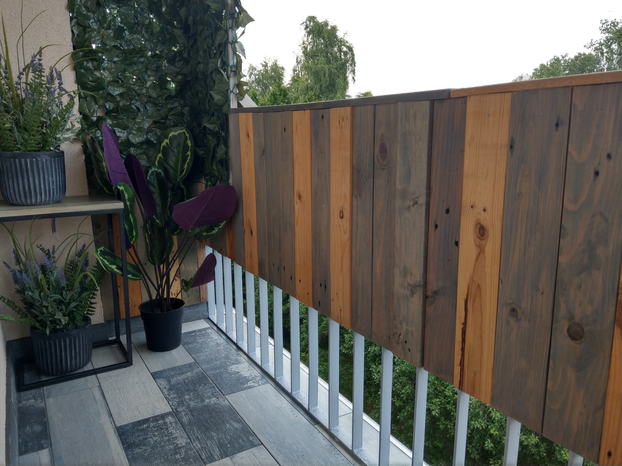 Balkon z drewnianą balustradą z desek o różnej kolorystyce, uzupełniony o rośliny w donicach i sztuczną zieloną ścianę.