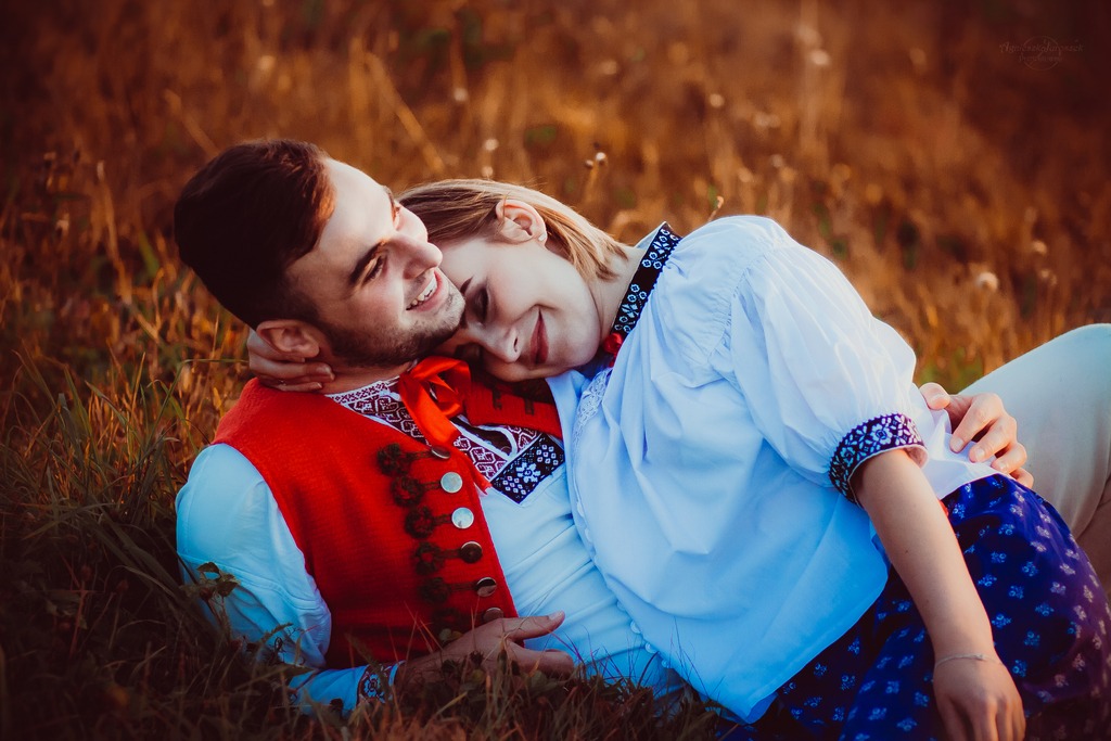 Uśmiechnięta para w strojach ludowych leży na trawie w Warszawie, mężczyzna w czerwonej kamizelce obejmuje kobietę w białej bluzce z haftem.
