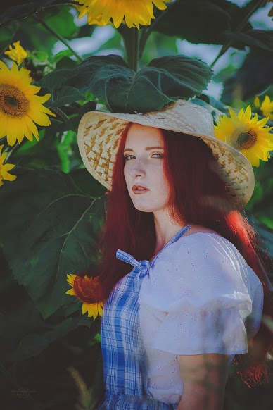 Portret kobiety z rudymi włosami w słomkowym kapeluszu, na tle pola słoneczników, ubrana w sukienkę w kratę z białymi rękawami.