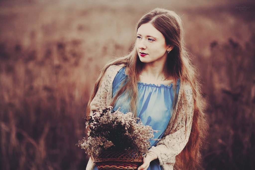Portret młodej kobiety z długimi, rudymi włosami, w błękitnej sukience i ażurowym swetrze, trzymającej wiklinowy koszyk z suszonymi kwiatami na rozmytym, brązowym tle.