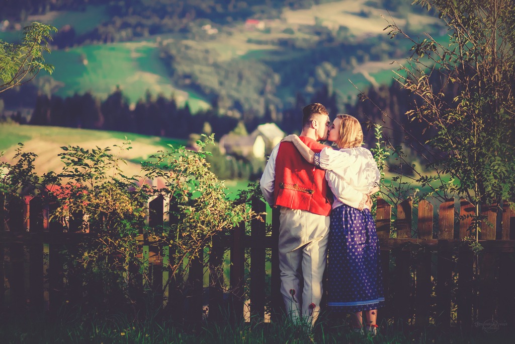 Zakochana para w strojach ludowych całuje się na tle malowniczego krajobrazu z drewnianym płotem.