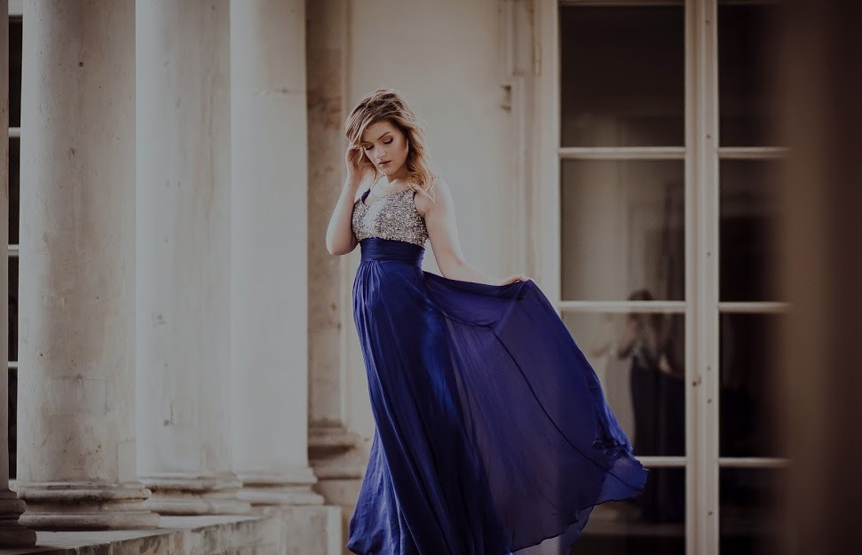 Kobieta w długiej, granatowej sukni z cekinową górą, pozująca przy kolumnach i oknach w stylu klasycystycznym, z delikatnym uśmiechem i ręką przy włosach.