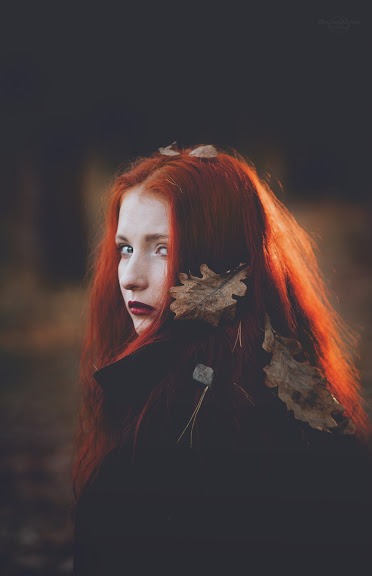 Portret kobiety z rudymi włosami, odwróconej przez ramię, z opadłymi liśćmi wplecionymi we włosy i ciemną szminką, w jesiennej scenerii.