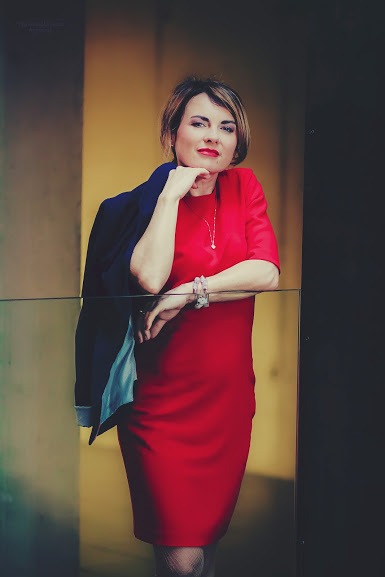 Portret kobiety w czerwonej sukience, opierającej się o szklaną balustradę, z marynarką przewieszoną przez ramię, na tle kolumn w ciepłych odcieniach.