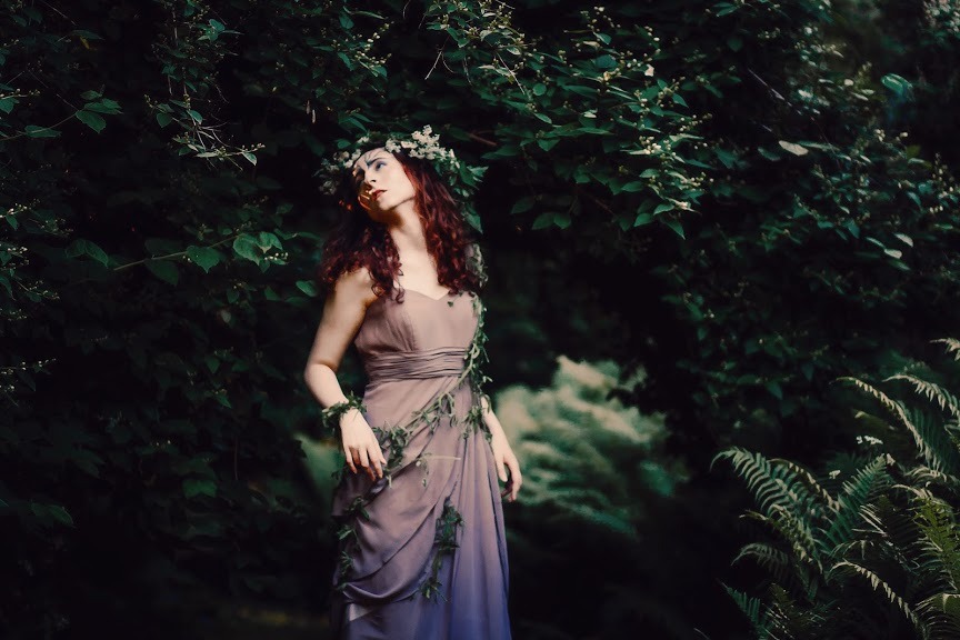Kobieta z rudymi kręconymi włosami w zwiewnej sukni i wianku z kwiatów, otoczona zielenią, pozująca w leśnej scenerii z paprociami.