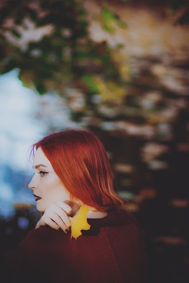 Portret kobiety z rudymi włosami w profilu, trzymającej żółty liść na ramieniu, ciemne usta, jesienna sceneria w tle.