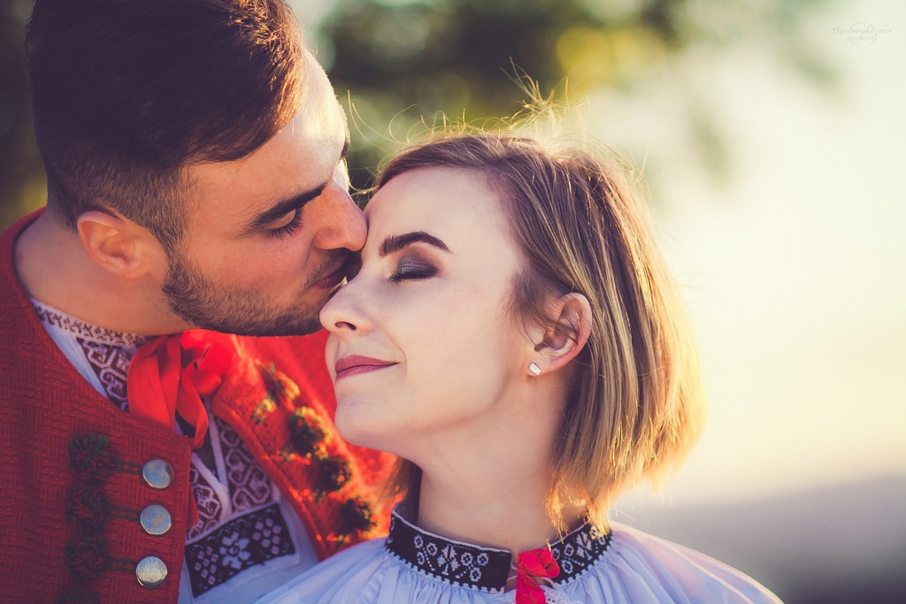 Mężczyzna w stroju ludowym całuje kobietę w czoło; oboje ubrani w tradycyjne stroje z haftem, ona ma zamknięte oczy i delikatny uśmiech.