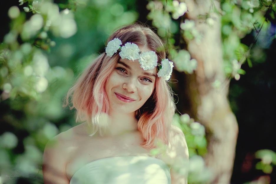 Portret uśmiechniętej kobiety z różowymi włosami i wiankiem z białych kwiatów na głowie, na tle kwitnących gałęzi drzew