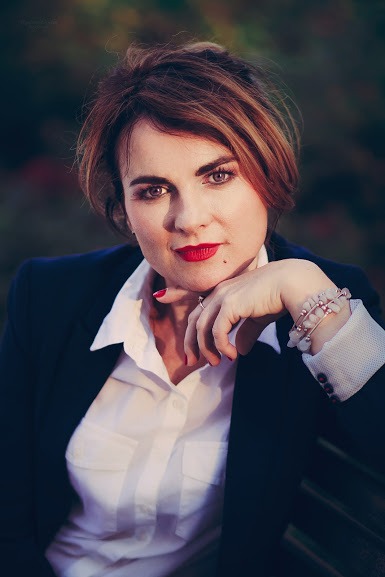 Portret kobiety w białej koszuli i granatowej marynarce, opierającej brodę na dłoni ozdobionej bransoletką z jasnych kamieni, z mocnym makijażem ust w odcieniu czerwieni, na rozmytym tle zieleni.