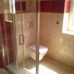 Nowoczesna kabina prysznicowa z transparentnego szkła w łazience z beżowo-czerwonymi płytkami, widoczny podwieszany sedes i okno w tle.