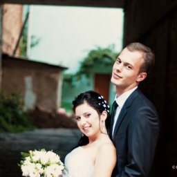 PrettyLook.pl - Uśmiechnięta para młoda w wąskim przejściu między budynkami, panna młoda z bukietem białych róż, pan młody w ciemnym garniturze i zielonym krawacie.