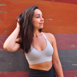 PrettyLook.pl - Portret kobiety w sportowym topie na tle malowanej ściany w odcieniach pomarańczy, czerwieni i szarości, z ręką we włosach i delikatnym uśmiechem.