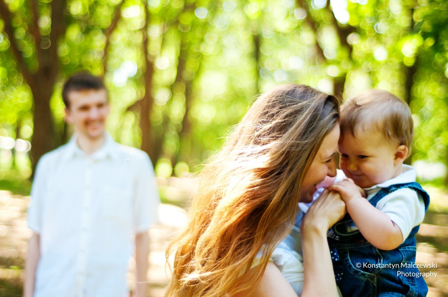 Matka trzymająca małe dziecko w dżinsowych ogrodniczkach na tle rozmytego parku i uśmiechniętego mężczyzny.