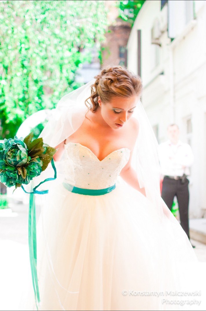 Panna młoda w białej sukni bez ramiączek z turkusową szarfą i bukietem turkusowych kwiatów, w tle mężczyzna w garniturze i jasny budynek.