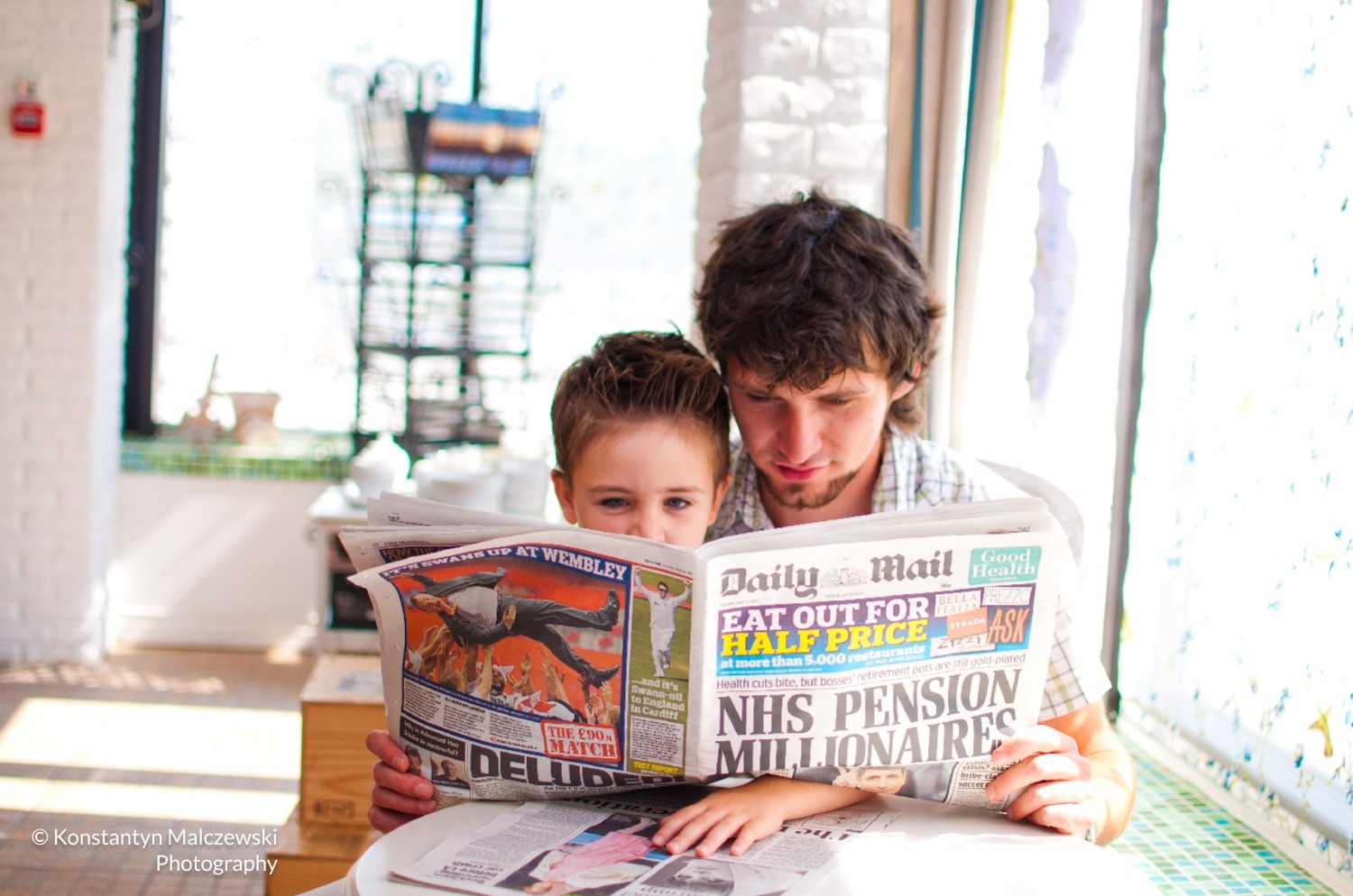 Ojciec i syn wspólnie czytają gazetę Daily Mail przy oknie, jasne wnętrze z ceglaną ścianą w tle.