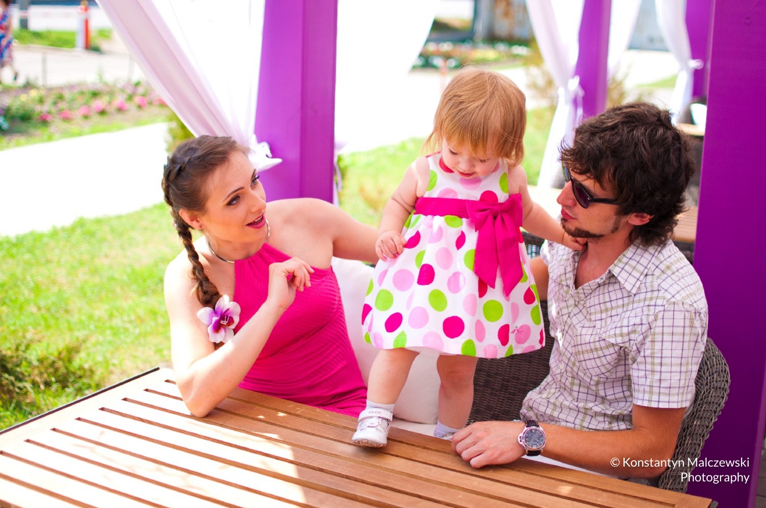 Uśmiechnięta matka w różowej sukience z kwiatem we włosach, ojciec w okularach i koszuli w kratę oraz mała dziewczynka w sukience w grochy stojąca na drewnianym stole w plenerze.