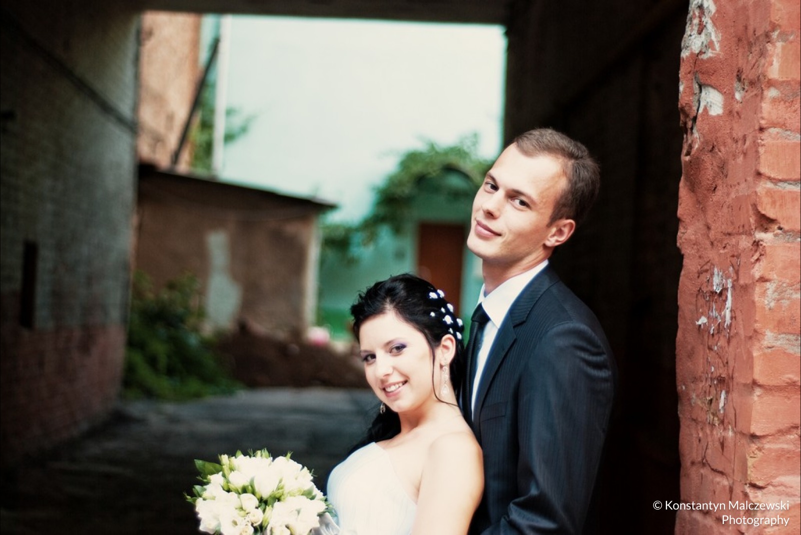 Uśmiechnięta para młoda w wąskim przejściu między budynkami, panna młoda z bukietem białych róż, pan młody w ciemnym garniturze i zielonym krawacie.