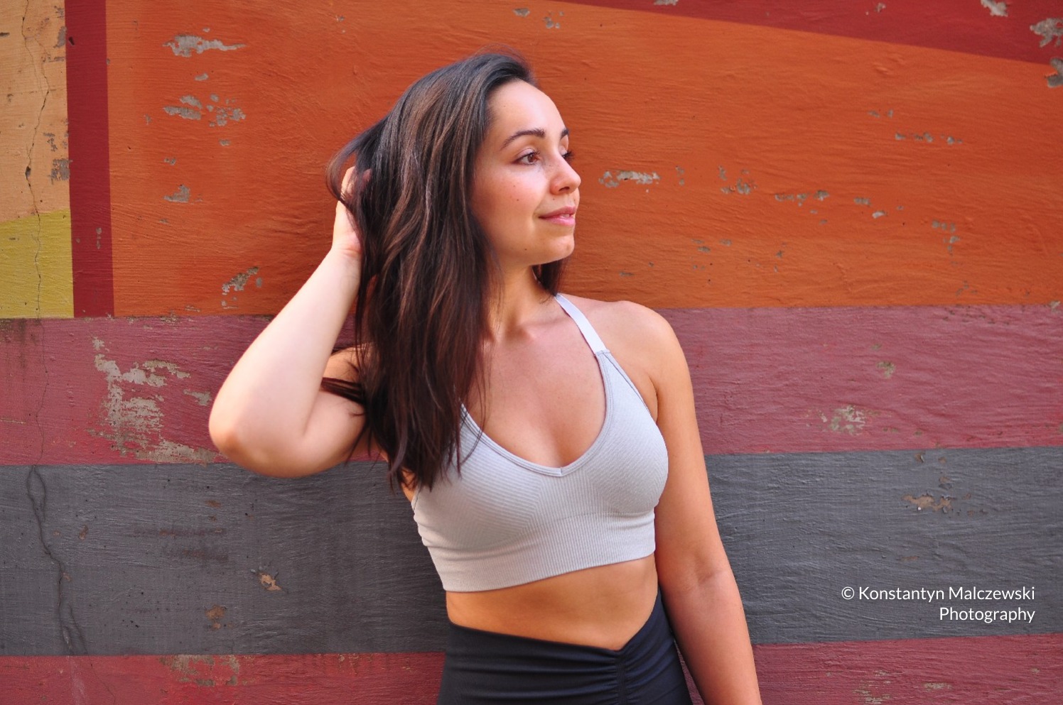 Portret kobiety w sportowym topie na tle malowanej ściany w odcieniach pomarańczy, czerwieni i szarości, z ręką we włosach i delikatnym uśmiechem.