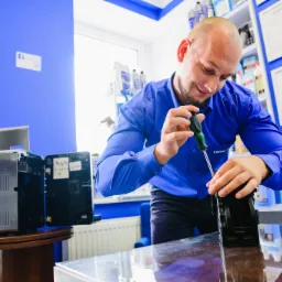 Mężczyzna w niebieskiej koszuli naprawia ekspres do kawy przy użyciu śrubokręta w pomieszczeniu z niebieskimi ścianami i półkami z akcesoriami.