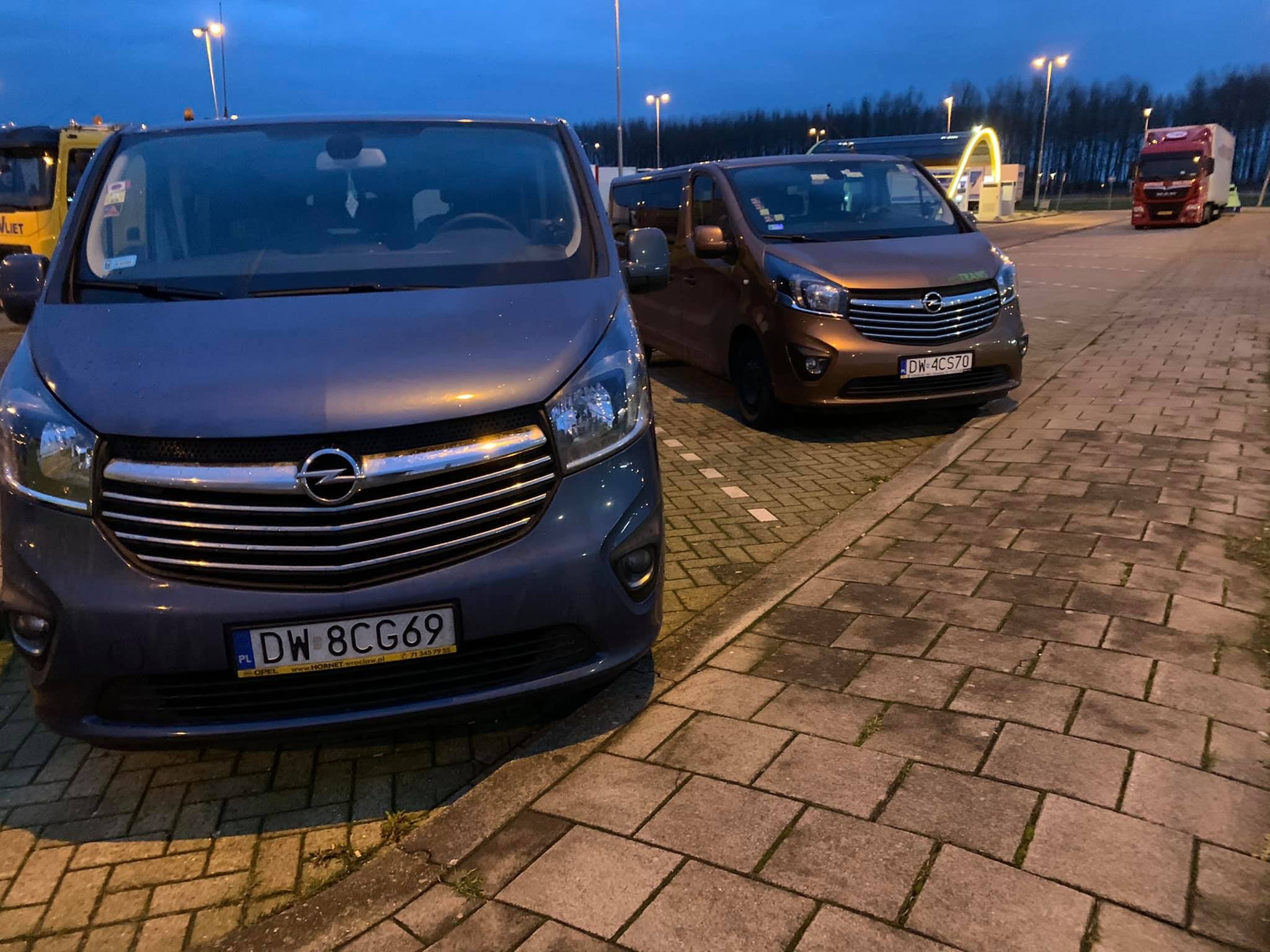 Dwa busy Opel Vivaro zaparkowane na parkingu przy stacji benzynowej, jeden szary, drugi brązowy, z widocznymi tablicami rejestracyjnymi DW 8CG69 i DW 4CS70.