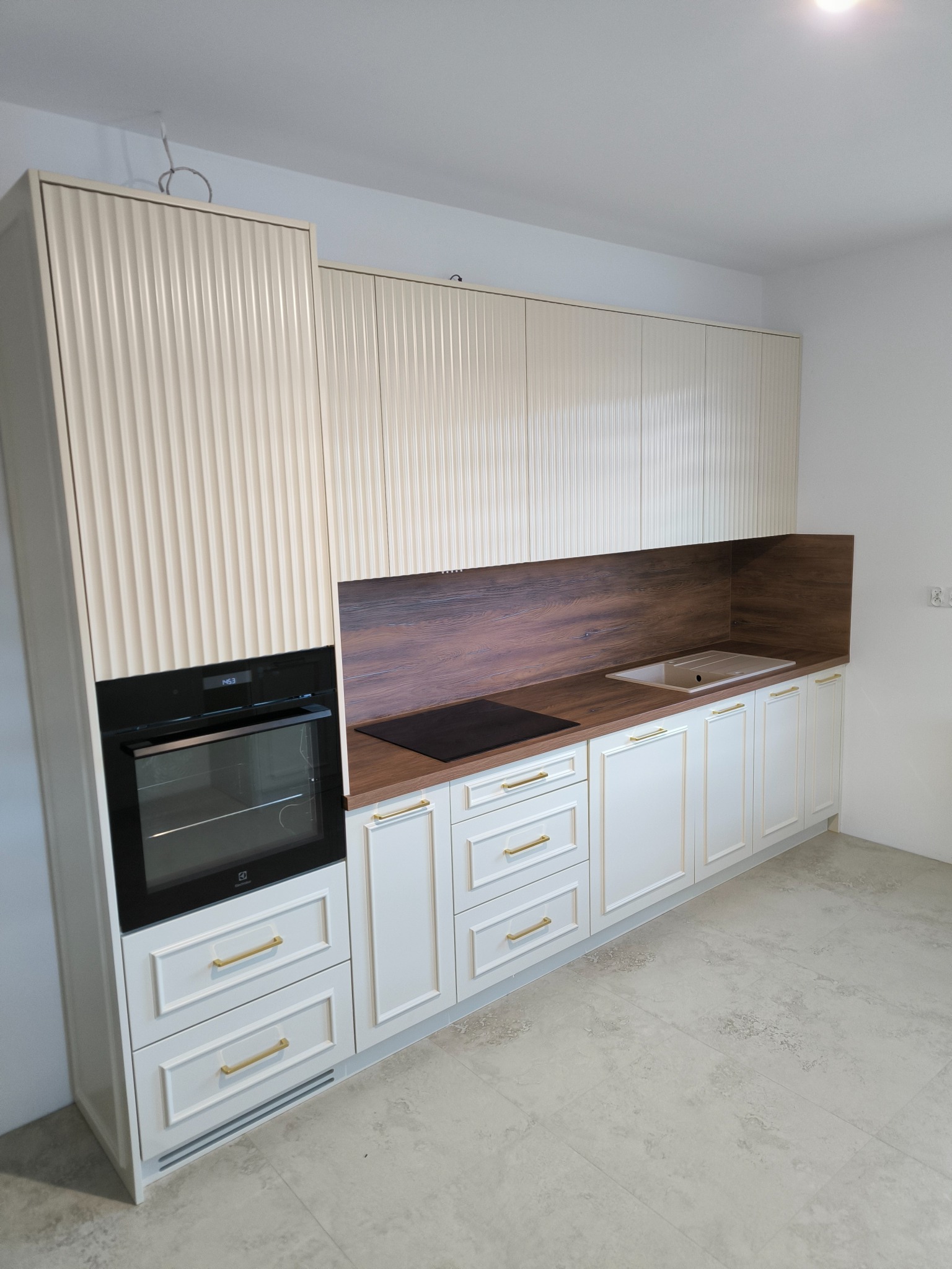 Nowoczesna kuchnia na wymiar z frezowanymi frontami w kolorze kremowym, złotymi uchwytami i drewnianym blatem, wbudowany piekarnik i płyta indukcyjna.