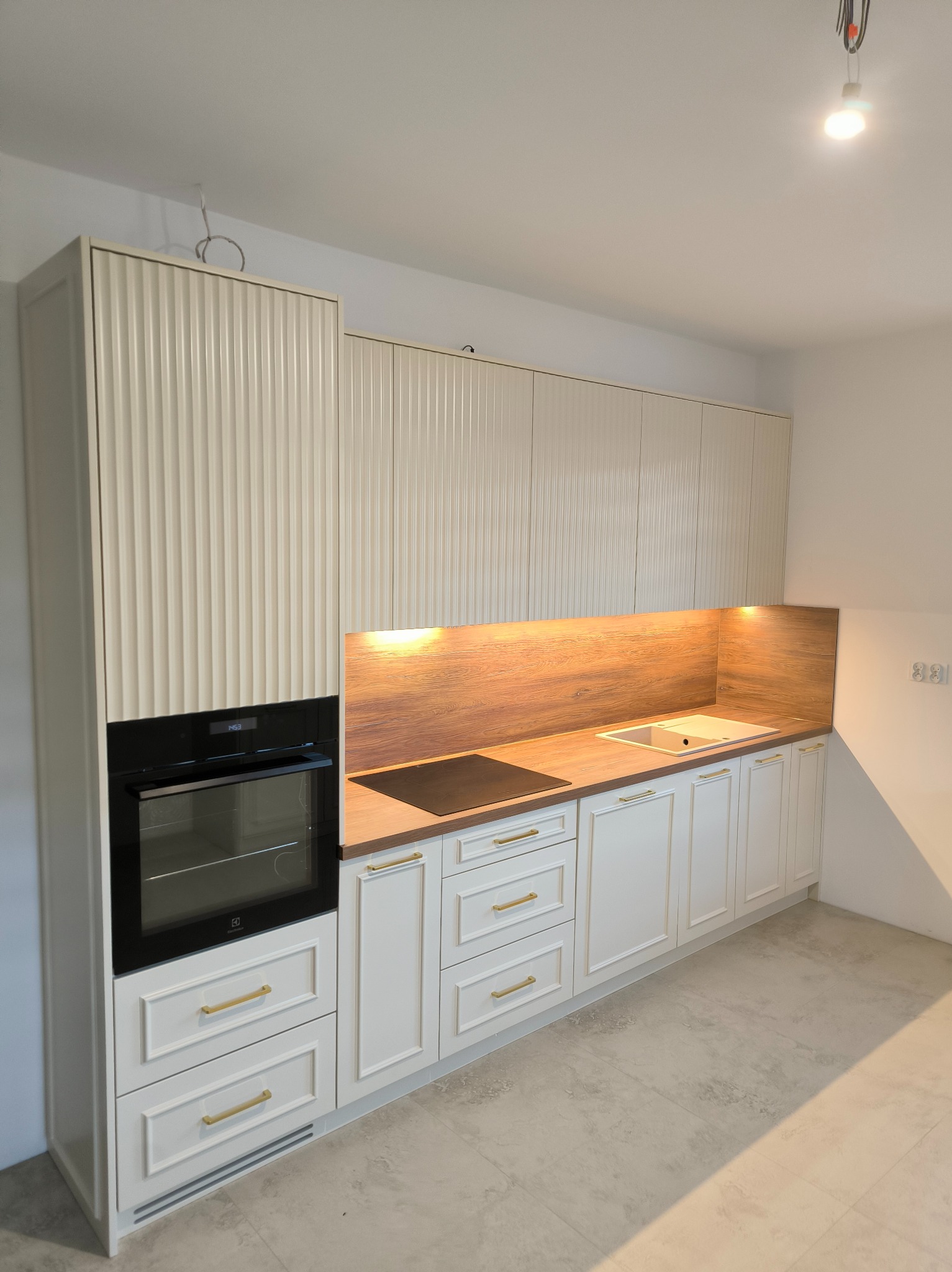 Nowoczesna kuchnia z frezowanymi frontami w odcieniach bieli i drewna, złote uchwyty, wbudowany piekarnik Electrolux, podświetlany blat i płyta indukcyjna.