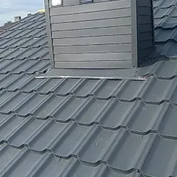 Szary dach z blachy trapezowej z nowo zamontowanym, obudowanym kominem z trzema kratkami wentylacyjnymi i daszkiem, widoczne fragmenty więźby dachowej w tle.