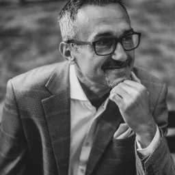 Czarno-białe portretowe ujęcie mężczyzny w okularach i marynarce w kratę, z ręką podpartą pod brodą, sugerujące zamyślenie.