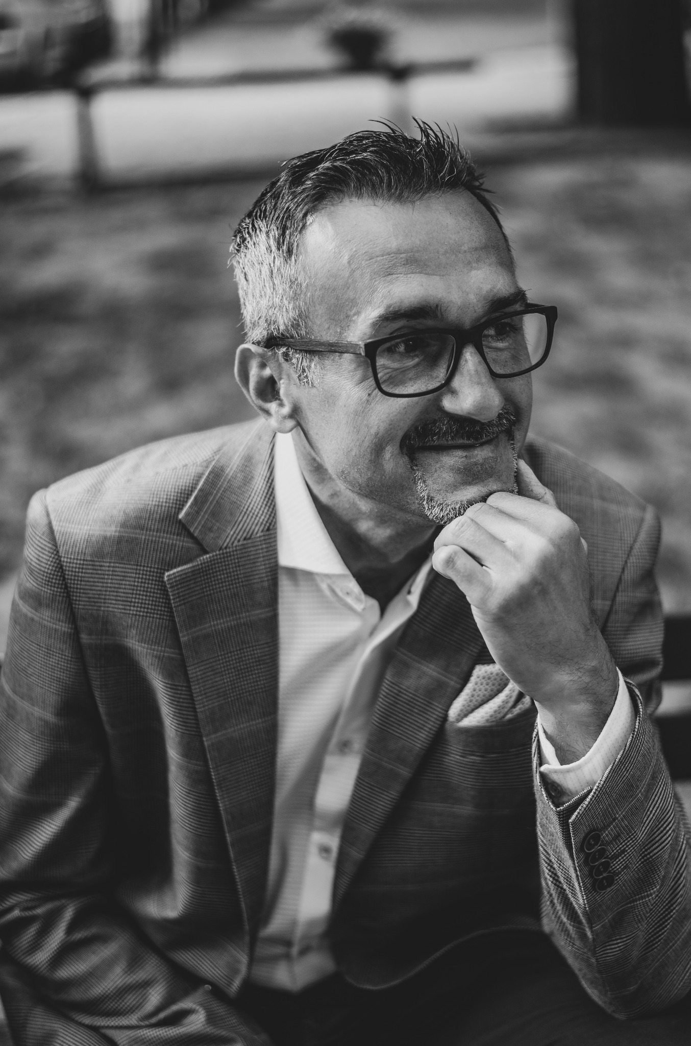 Czarno-białe portretowe ujęcie mężczyzny w okularach i marynarce w kratę, z ręką podpartą pod brodą, sugerujące zamyślenie.