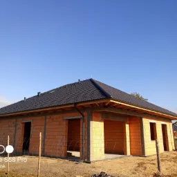 Niedokończony budynek jednorodzinny z cegły z ciemnym dachem i widoczną drewnianą konstrukcją podbitki dachowej, sfotografowany w słoneczny dzień.