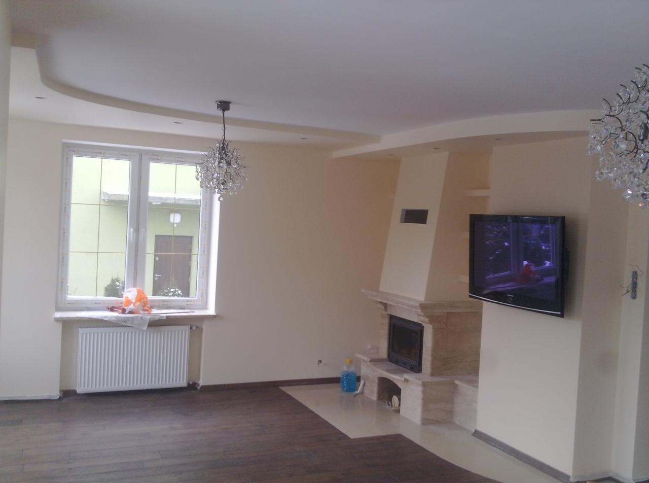 Salon po remoncie z kremowymi ścianami, podwieszanym sufitem z dekoracyjnym oświetleniem, marmurowym kominkiem i telewizorem na ścianie.