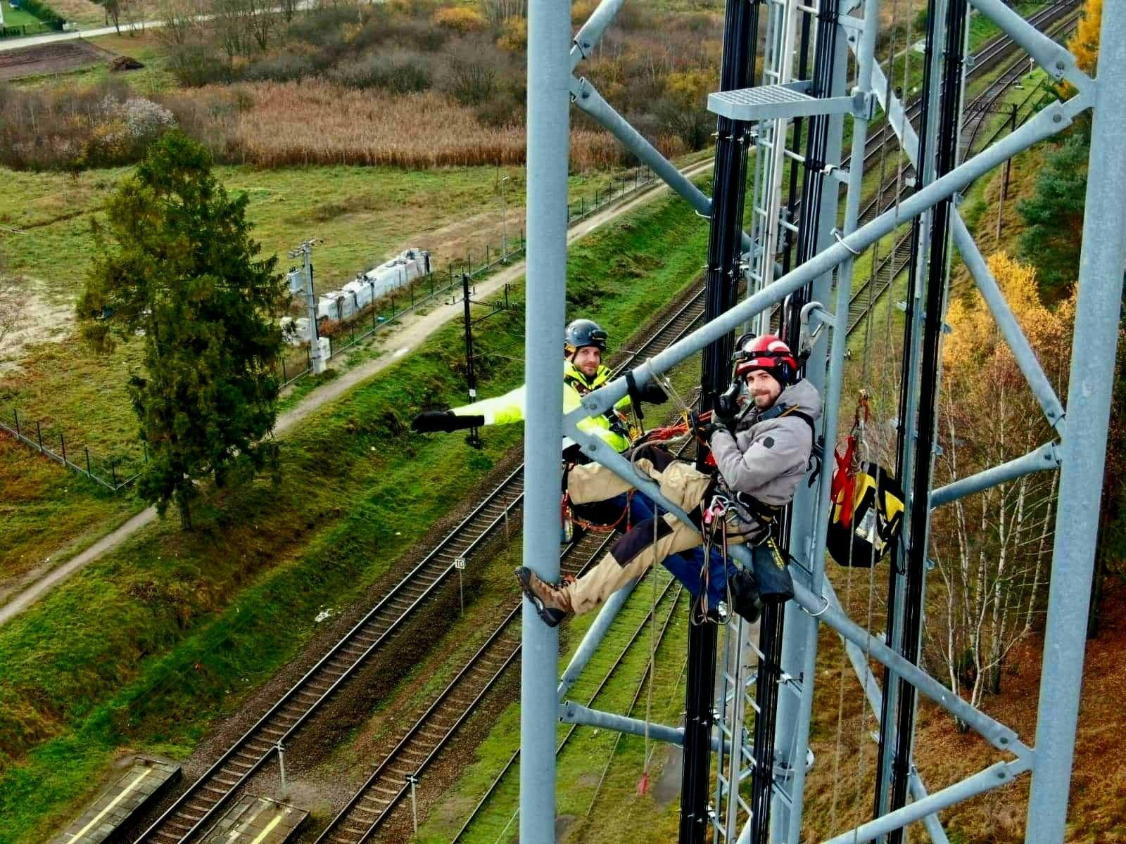 Dwóch pracowników w kaskach, zabezpieczeniach i odzieży roboczej, na wysokości słupa telekomunikacyjnego w Gdyni, z widokiem na tory kolejowe i zielone tereny.