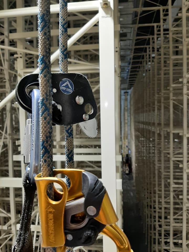 Sprzęt do prac wysokościowych: lina, karabinek, przyrząd zaciskowy Ascension, widok na metalową konstrukcję magazynową w tle.