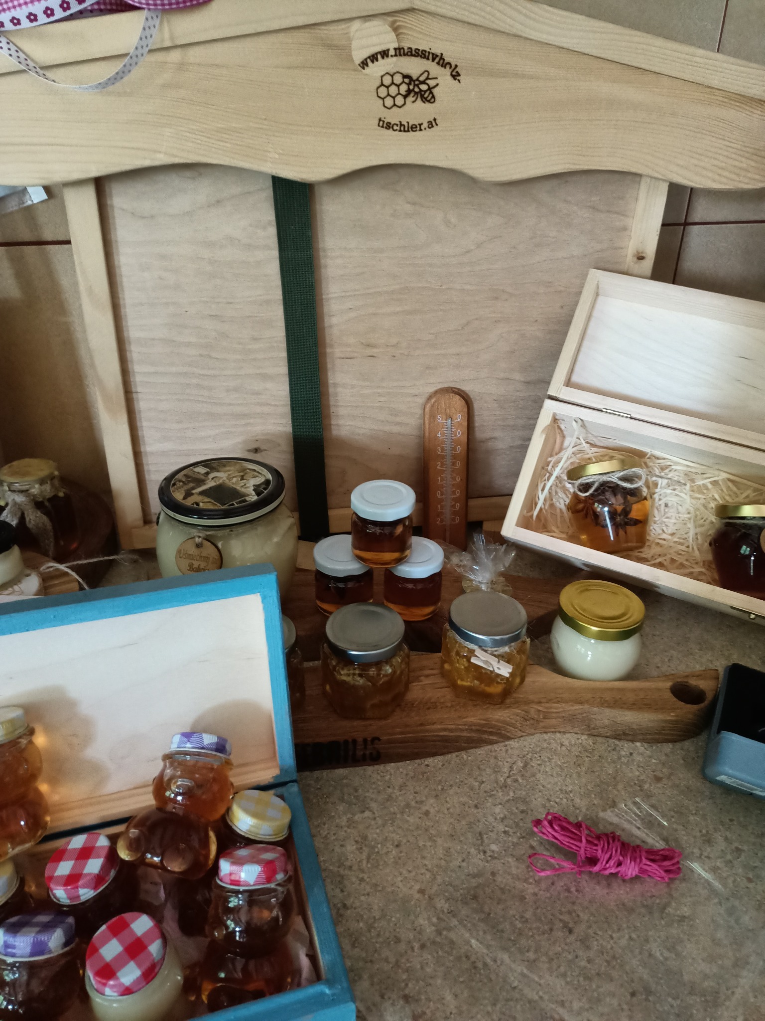 Różnorodność miodów w szklanych słoikach o różnych kształtach i wielkościach, niektóre w drewnianych pudełkach, prezentowane na drewnianej desce i w drewnianej konstrukcji z logo pszczelarza.