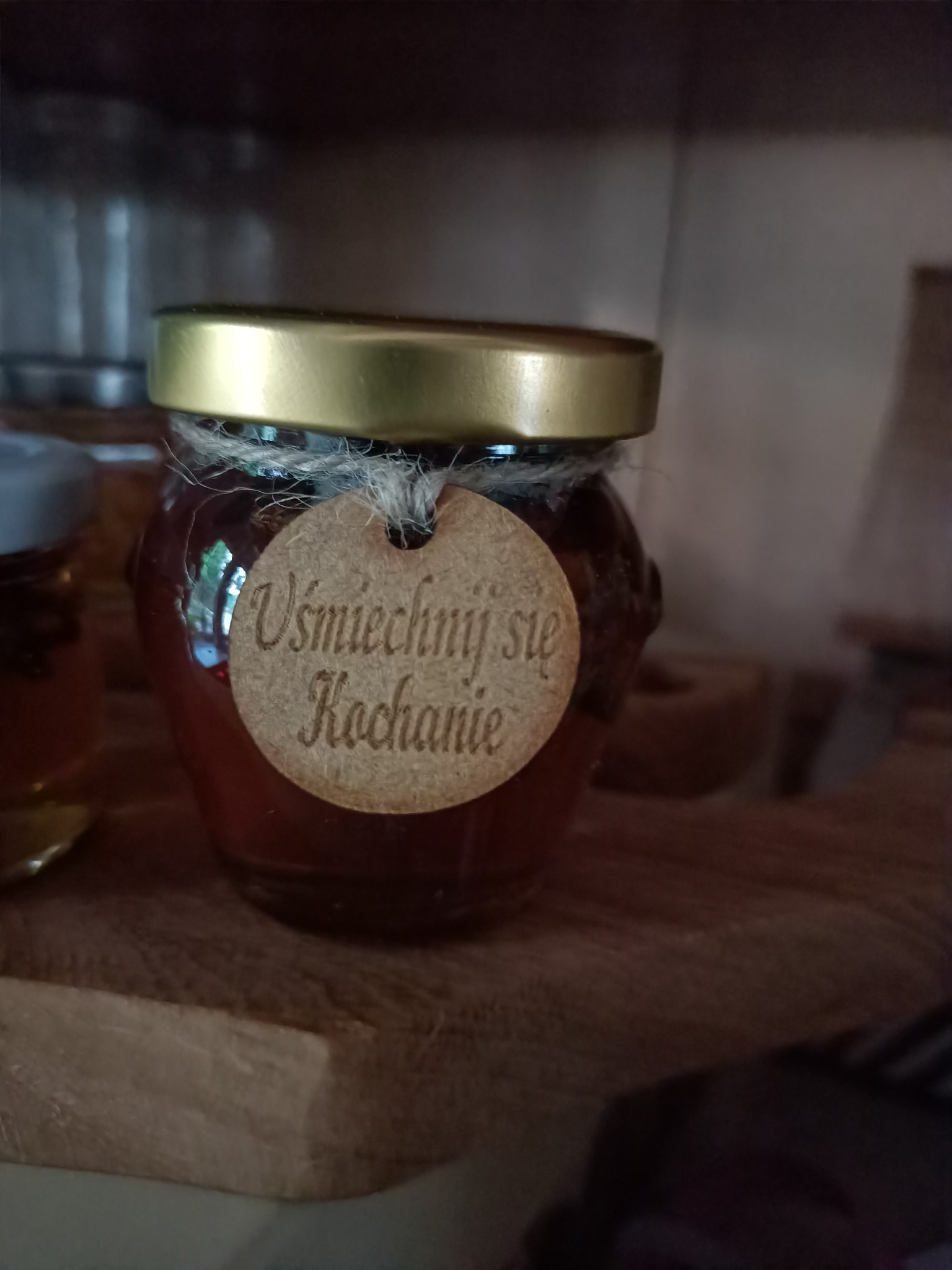 Słoik z zawartością o bursztynowej barwie, zamknięty złotym wieczkiem i ozdobiony jutowym sznurkiem oraz drewnianą etykietą z napisem 'Uśmiechnij się Kochanie'.