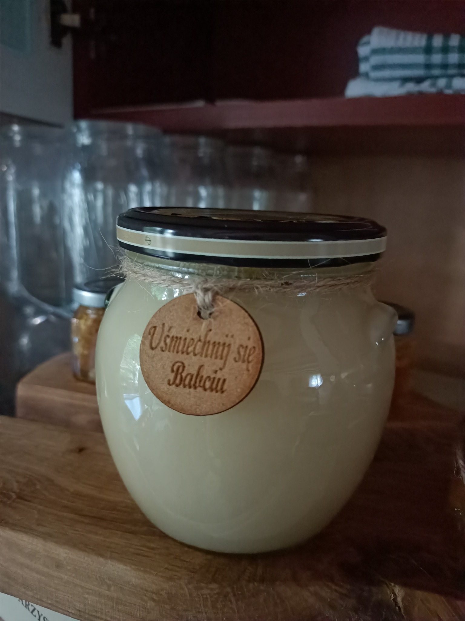 Słoik z kremowym produktem, prawdopodobnie spożywczym, z etykietą 'Uśmiechnij się Babciu', owinięty sznurkiem, stojący na drewnianej półce w otoczeniu innych słoików i naczyń w szafce.