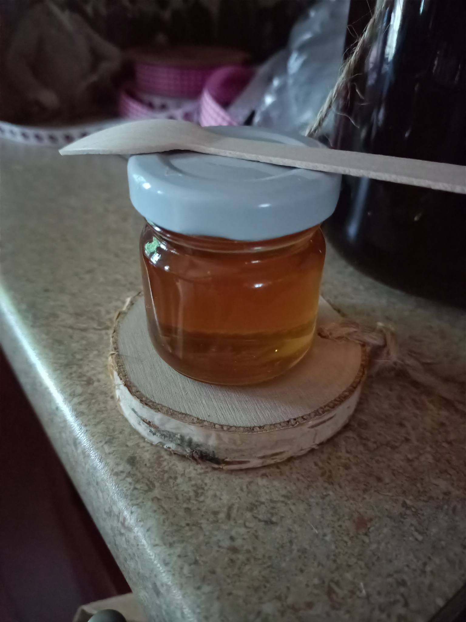 Mały słoiczek z bursztynowym płynem, zamknięty jasnoszarą nakrętką, leży na drewnianym plastrze z korą, z drewnianą łyżeczką na wierzchu, w tle widoczny słoik z ciemnym płynem i dekoracyjna wstążka.