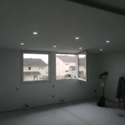 Wnętrze pokoju po montażu oświetlenia punktowego w suficie, widoczne dwa okna z widokiem na budynki mieszkalne, ściany przygotowane do malowania, na podłodze widoczne ślady po pracach remontowych...