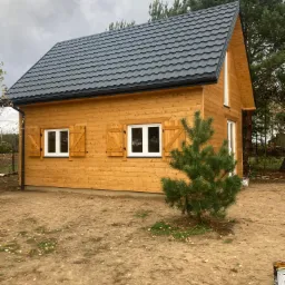 Gotowy, parterowy dom szkieletowy z ciemną blachodachówką, drewnianymi okiennicami i małą sosną przed budynkiem, widok z boku.