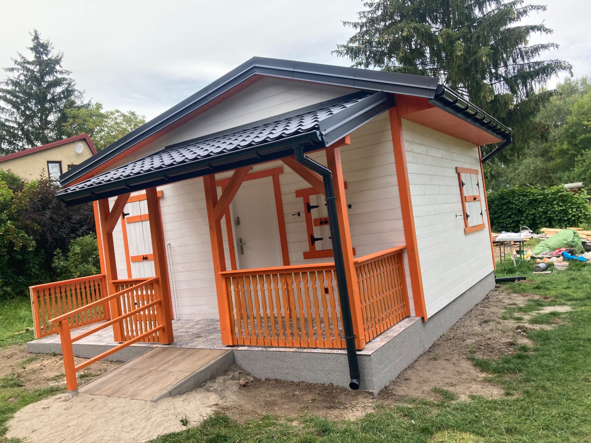 Biały budynek z pomarańczowymi akcentami, czarnym dachem i rynnami, otoczony zielenią, z rampą wejściową.